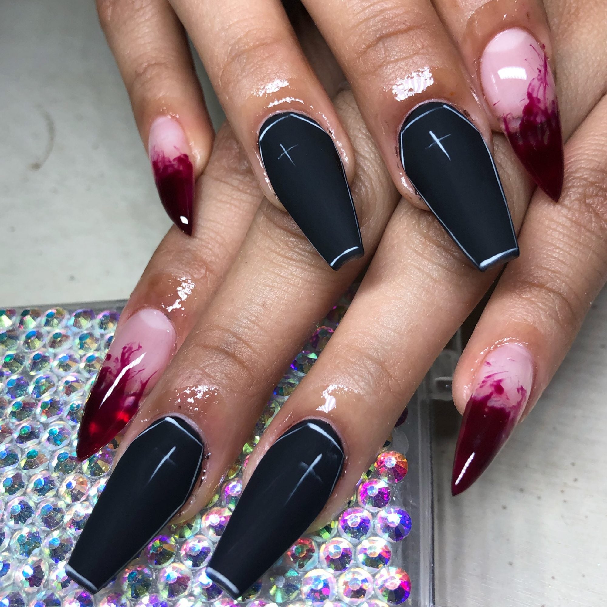 A set of goth nails where the outer 2 fingers have sharp clear faux nails that have swirling, blood-red tips and the inner two are matte-black and long, coffin-shaped with a single white cross decorating the nail bed. 