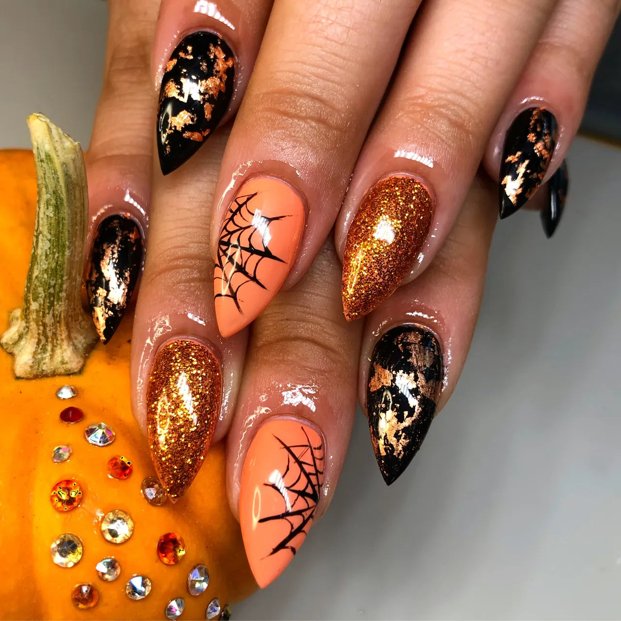 A halloween-theme faux nail set that has black and gold marbled nails on the outside and one orange gold glitter nail and one glossy orange nail with black spiderweb art. The hands that are modeling the nails are a light carmel complexion and resting on a bejeweled pumpkin.