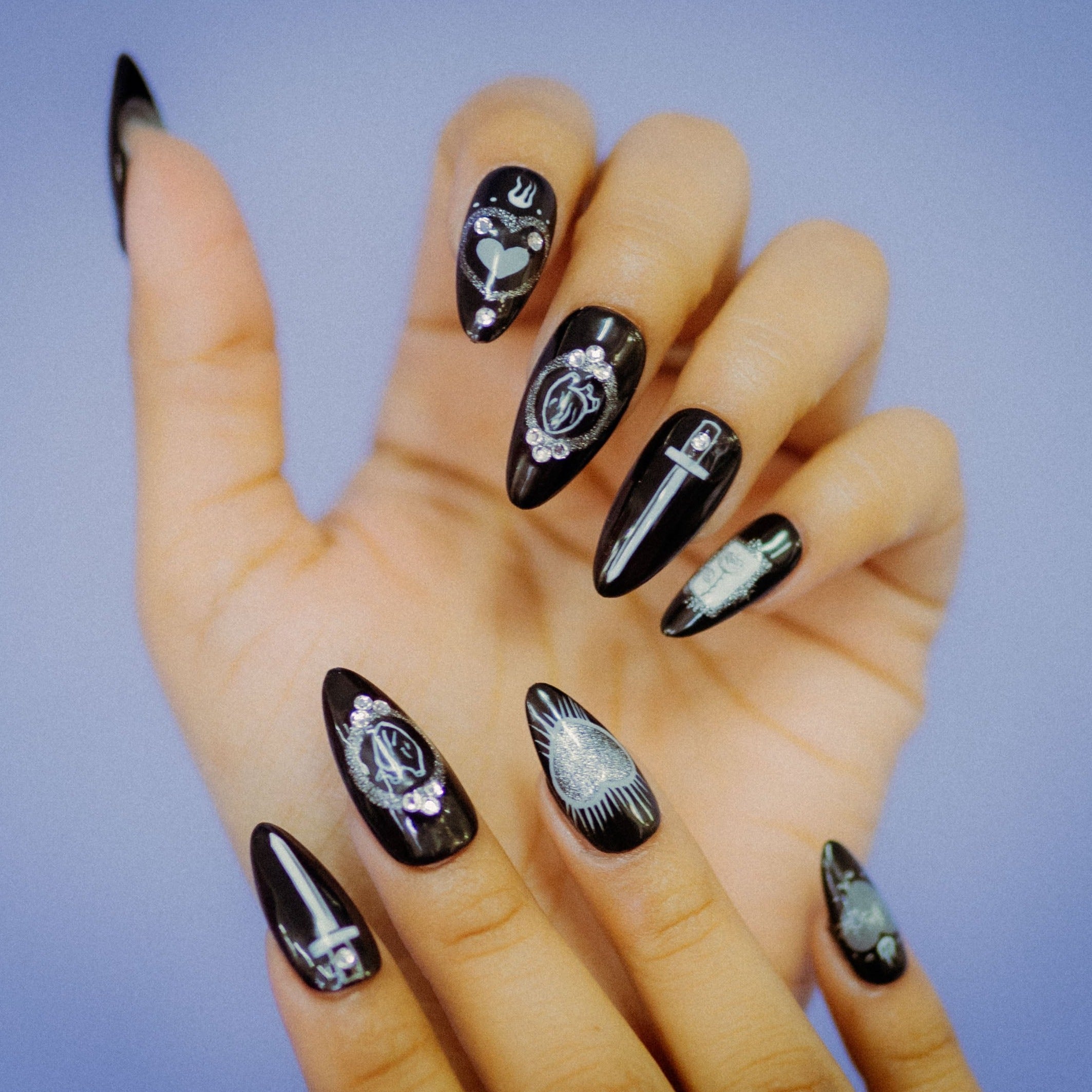 Light-skinned hands showing off the Mea Culpa faux nails, each nail is black, with white designs that are embellished by rainbow holographic details. The nails incorporate Mexican, Catholic, and witchy imagery, such as blades, hearts, and flowers, to create a uniquely gothic aesthetic